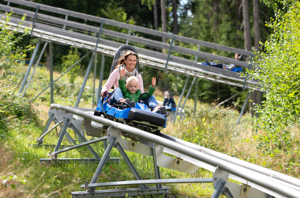 Eine Frau fährt mit einem Kind in einem Schlitten auf einer Sommerrodelbahn bergab. Beide heben die Arme, im Hintergrund verläuft die Schienenstrecke durch grünen Wald.