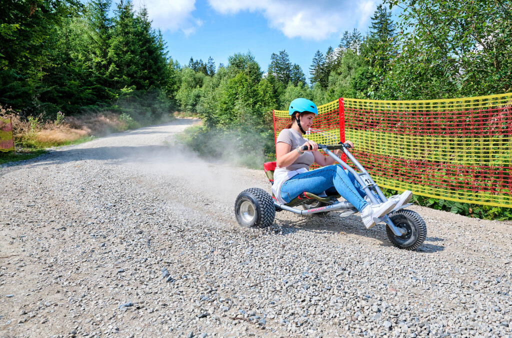 Eine Frau mit Helm fährt mit einem Mountaincart eine Schotterstrecke bergab. Staub wirbelt auf, neben der Strecke verläuft ein Sicherheitsnetz, umgeben von Wald.