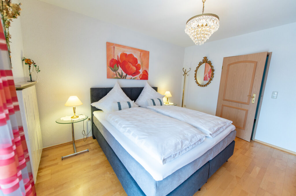 Doppelbett mit weißen Kissen und Decken, zwei Nachttische mit Lampen, Wandbild über dem Bett und Zimmertür im Hintergrund.