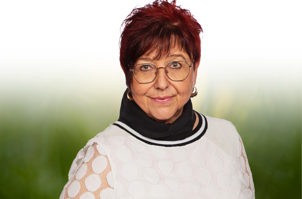 Porträt der Gastgeberin Ute Vogl mit kurzen roten Haaren und Brille. Sie trägt ein helles Oberteil und blickt freundlich in die Kamera vor unscharfem grünem Hintergrund.